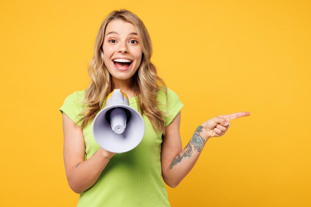 Young female direct sales representative stands smiling holding a megaphone wearing a green shirt set against a yellow background